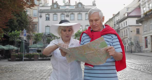 Senior Male and Female Tourists Walking with a Map in Hands Looking for Route