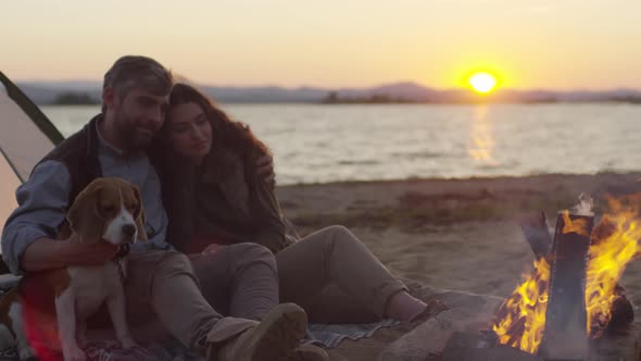 Romantic Couple Sitting with Dog by Campfire at Beach