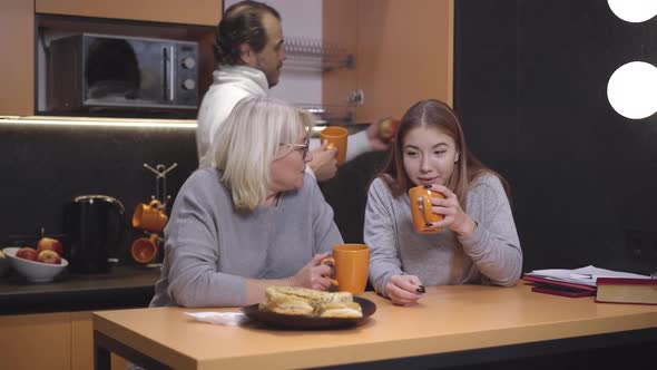 Positive Mother Drinking Tea with Teenage Daughter in Kitchen As Father Taking Cup From Locker at
