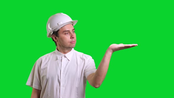 Young Male Smiling Engineer in White Hardhat and Shirt Standing and Showing Hand Palm on Green