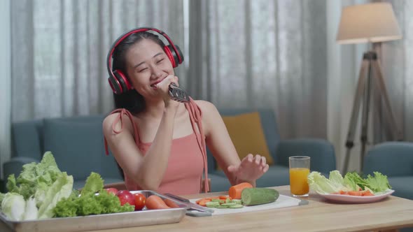Asian Woman Listening To Music With Headphones And Singing While Slicing Carrot At Home