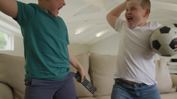 Happy caucasian boy with brother watching tv and holding football in living room