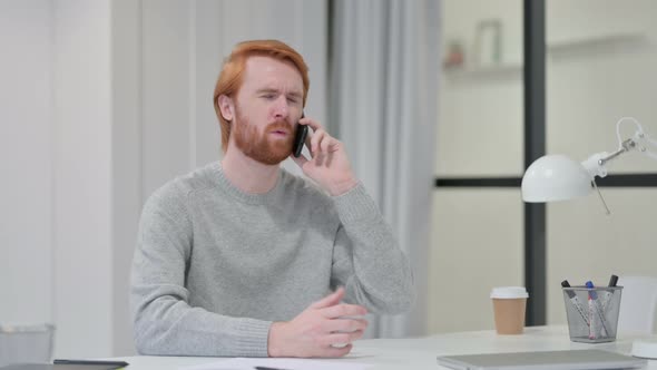 Angry Young Redhead Man Talking on Smartphone at Work 