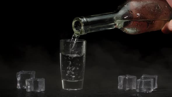 Barman Hand Pours Vodka Tequila or Sake From Bottle Into Glass on Black Background with Ice Cubes