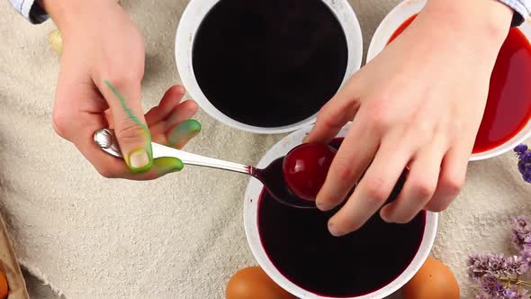 Caucasian Child Paints an Easter Egg with a Metal Spoon and Dips It Into a White Bowl with Red Dye