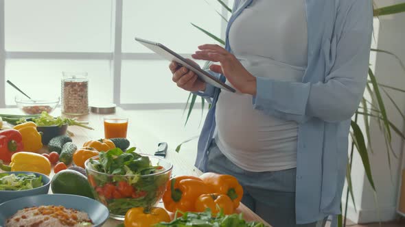 A Pregnant Woman Using a Tablet Prepares Healthy Food in a Bright Kitchen