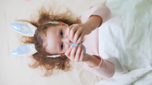 Happy Baby Girl with a Rim of Bunny Ears on Her Head Lies on Her Back on White Bed at Home in the