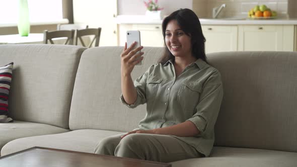 Young Indian Woman Sitting on Sofa Video Calling in Mobile Phone App at Home