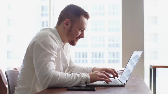 Side View of Cheerful Bearded Young Businessman Working on Laptop at the Desk