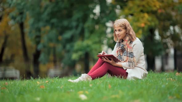 Young Woman Using Tablet Outdoor Sitting on Grass and Smiling