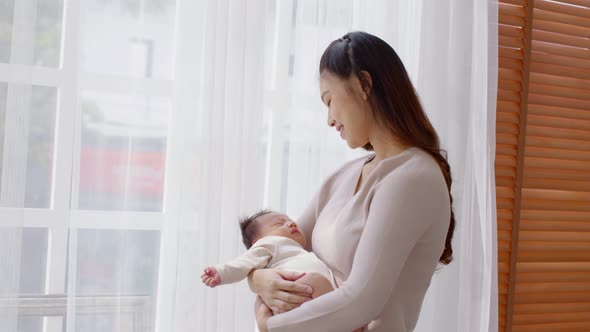 Beautiful Asian woman holding newborn baby in her arms standing in front of windows