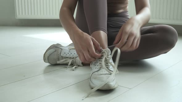 Young Girl in Sportswear Sits on Floor on Knees Untying Shoelaces on Sneakers