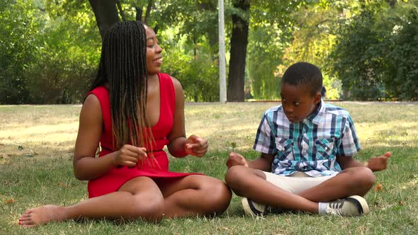 A Young Black Mother and Her Son Sit on Grass in a Park and Celebrate