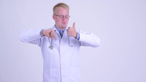 Confused Young Blonde Man Doctor Choosing Between Thumbs Up and Thumbs Down