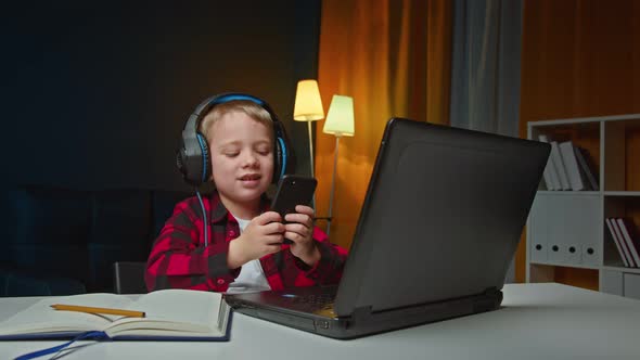 Cheerful Energetic Little Boy Using Smartphone and Laptop and Listening to Music in Headphones at