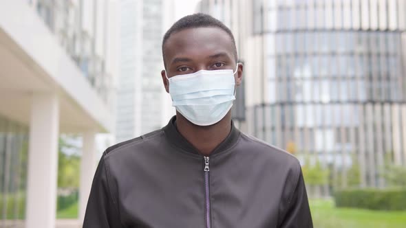 A Young Black Man in a Face Mask Nods at the Camera - Office Buildings in the Blurry Background