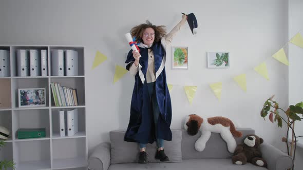 Graduation Celebrations Joyful Female Student in an Academic Mantle Has Fun Jumping on Couch with