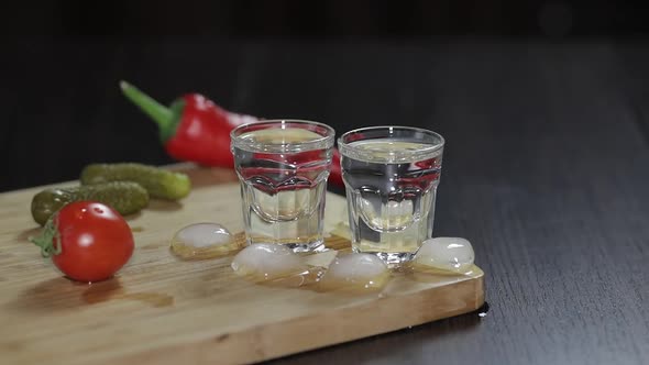 Vodka in Shot Glasses on Rustic Wood Board. Adding Ice Cubes