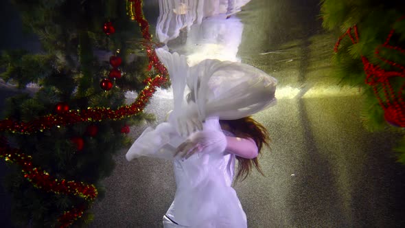 Beautiful Woman in White Dress and Fur Hat Is Swimming Underwater in Christmas Decorations