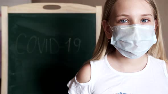 A Little Girl at Home Wears a Medical Mask Behind Which is the Inscription COVID19