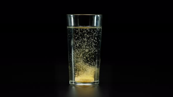 Closeup of Falling Yellow Fizzy Medicine Tablet Into Glass with Water on Black Background Shooting