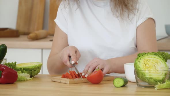 Young Blonde Woman with Curly Hair Slices a Red Tomato Sitting at a Table in the Kitchen