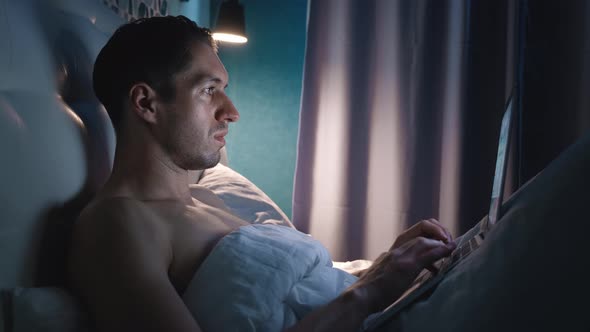 Portrait of Man Freelancer Lying on a Bed in the Bedroom at Night at Home and Working with Laptop