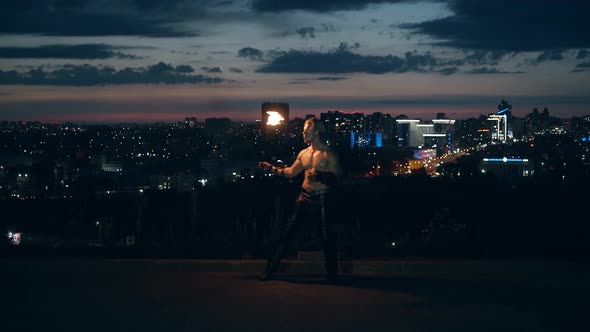 Young Blond Male Does Tricks with Fire Breaths Fire in the Middle of the Night with City Skyline