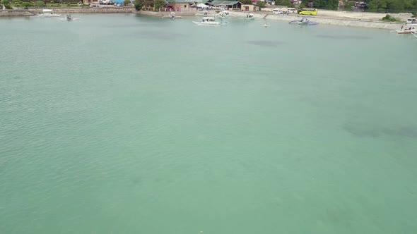 Low drone flying fast over shallow waters of Badian Bay tilting up to reveal the port and town of Mo