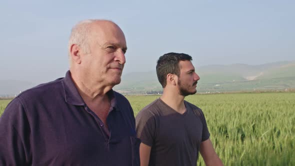 Old farmer walking with a young farmer in a green wheat field