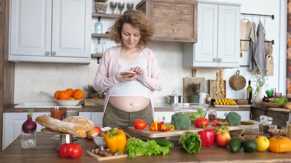 Beautiful Pregnant Female Using Smartphone And Cooking On Kitchen.