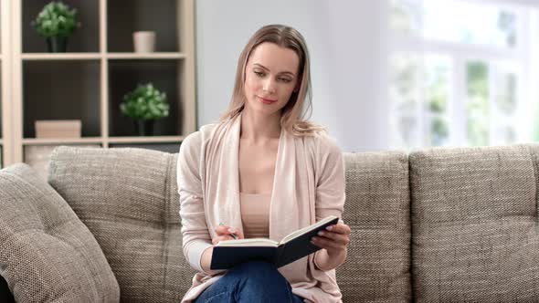 Pleasant Young Blonde Woman Thinking Writing Notes in Diary or Making to Do List Relaxing on Couch