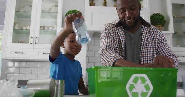 Happy african american father and son standing in kitchen putting plastic rubbish in recycling box