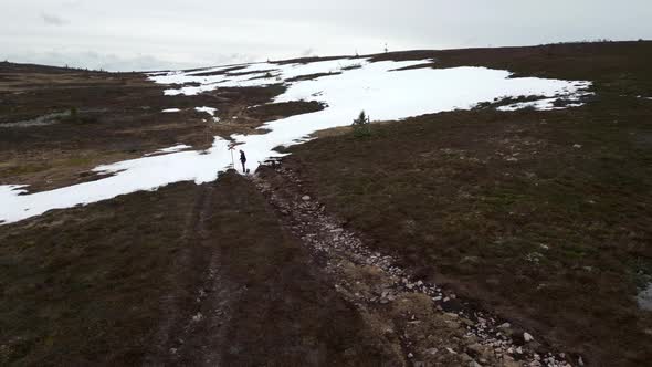 A woman and her french bulldog walking on a mountain gravel path with snow infront of them. Aerial f