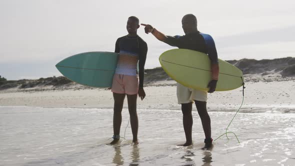 African american father and teenage son standing on a beach holding surfboards and talking