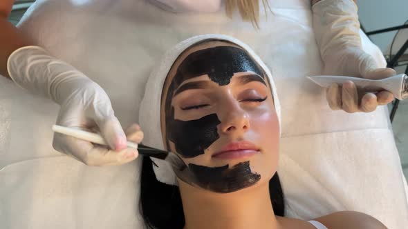 Cosmetologist Applying Black Mask on Pretty Woman Face Wearing Black Gloves Gorgeous Woman in Spa