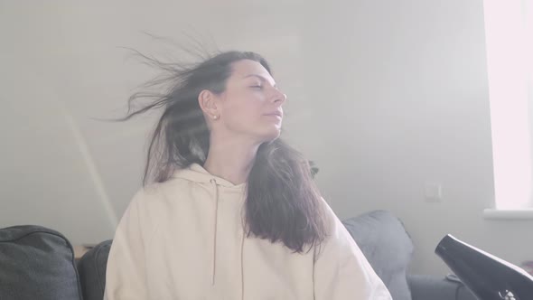 Young Millennial Caucasian White Woman Drying Hair with Hairdryer at Home on the Sofa in Living Room