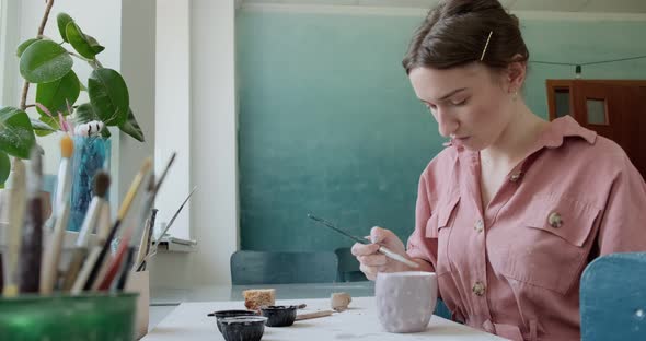 Female Potter Sitting and Stirs Paint with a Brush a Cup on the Table. Woman Making Ceramic Item