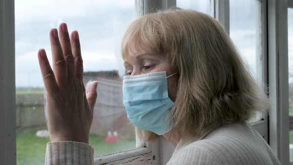 Sad Mature Woman In Medical Mask Looks Out At Street With Her Hand On Glass