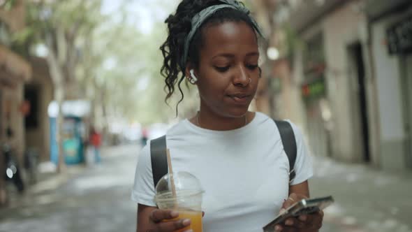 Cheerful brunette African woman texting on mobile and drinking juice