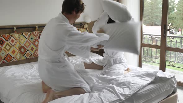 A Cheerful and Active Married Couple in White Coats Fights with Pillows on a Soft Bed in a Hotel