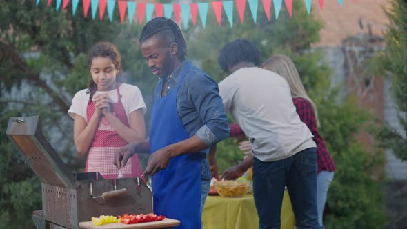 Teenage Girl and Adult Man Cooking Barbecue Grill Outdoors with Friends Setting Table at Background