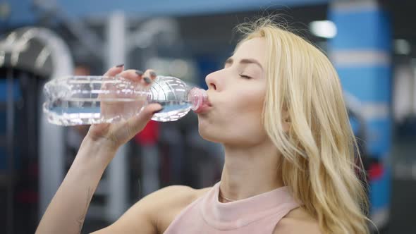 Headshot of Gorgeous Caucasian Blond Woman Drinking Water in Gym and Looking at Camera