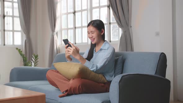 Asian Female Using Smartphone And Smiling In Living Room