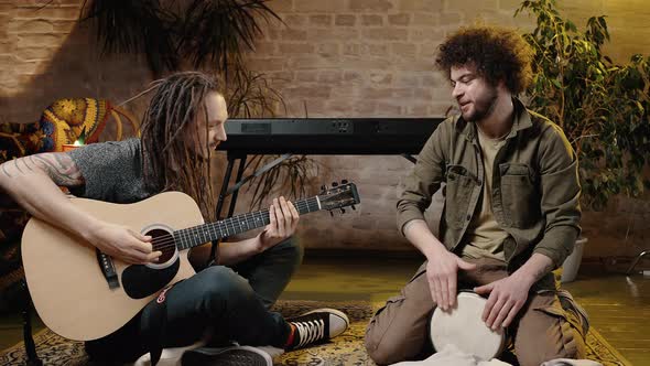 Two Adult Male Friends Performing Music Together Playing Guitar and Beating Drum Sitting on Floor