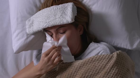 Sick Woman With Towel on Forehead Lying in Bed, Coughing and Blowing Nose, Flu