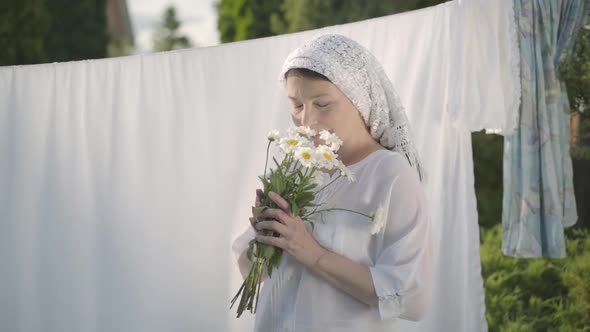 Cute Mature Woman with White Shawl on Her Head Tears Off Daisy Petals at the Clothesline Outdoors