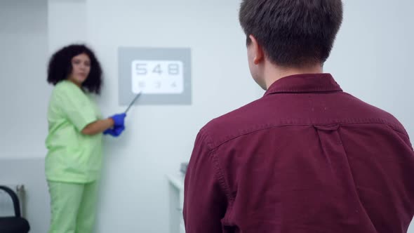 Shooting Over Shoulder of Caucasian Man Sitting in Ophthalmologic Clinic with Blurred Woman Pointing