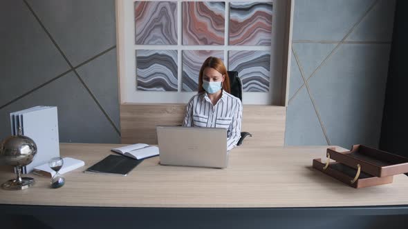 Young Woman Employee in a Medical Facial Mask Works in the Office at the Computer. 