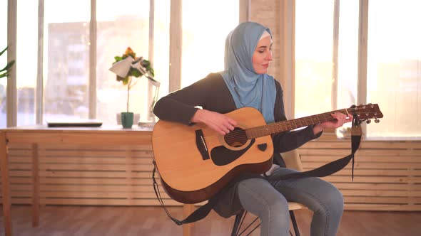 Young Muslim Woman Musician in Hijab Playing Guitar in Modern Apartment